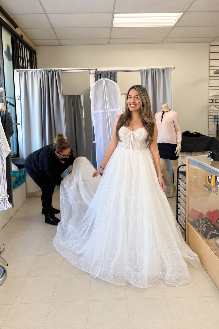 A bride in a sparkling ball gown while the seamstress adjusts the hem